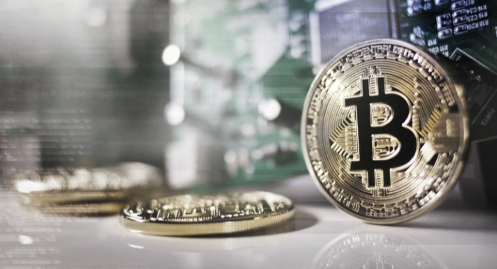 金色のビットコインコインがテーブル上に並び、手前に一枚が立てかけられ、奥には数枚が平置きされた状態で、背景にはぼかされた回路基板と光の反射が見える写真