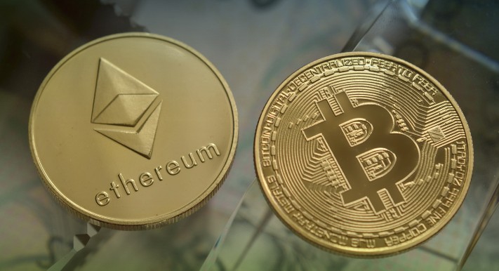 金色のEthereumコインとBitcoinコインが並んでおり、光沢のある反射面の上に置かれている写真